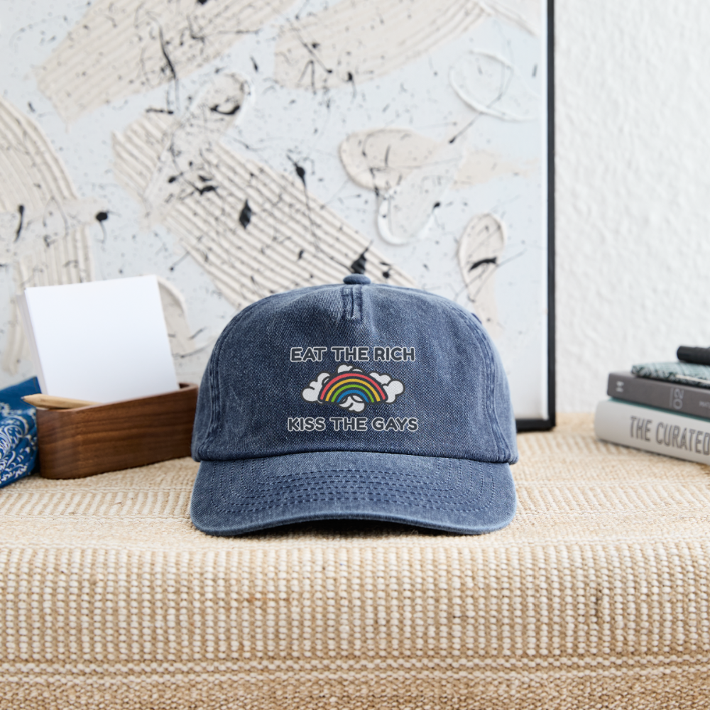 Die blaue Vintage Denim Cap mit dem Schriftzug „Eat The Rich Kiss The Gays“ liegt dekorativ auf einer geflochtenen Oberfläche. Das farbenfrohe Regenbogen-Motiv auf dem verwaschenen Stoff macht die Basecap zu einem starken Statement für sichtbaren Pride im Alltag.