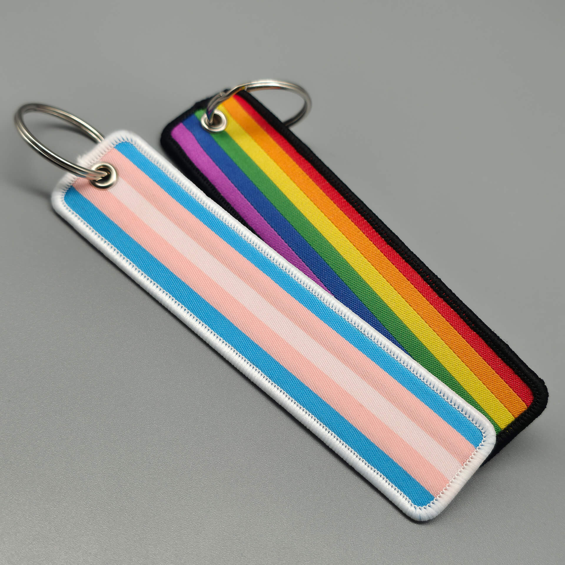Zwei flache Textil-Schlüsselanhänger liegen nebeneinander auf einem grauen Untergrund. Links zeigt das gewebte Band die pastellfarbenen Streifen der Trans-Flagge mit weißer Kettelnaht. Rechts leuchten die kräftigen Farben der Regenbogen-Flagge, eingerahmt von einer dichten schwarzen Naht. Ein robustes Pride-Accessoire für den täglichen Aktivismus.