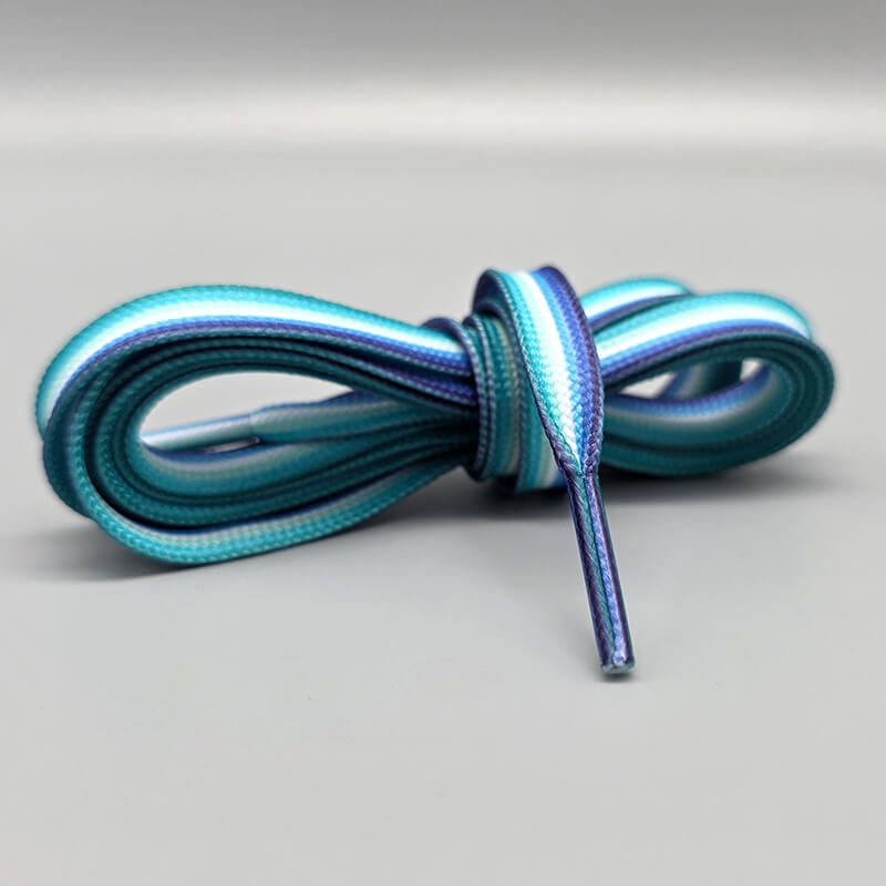Ein Paar zusammengerollte Schnürsenkel in den Farben der Gay Man- bzw. mlm-Flagge (man loves men). Die Schnürsenkel zeigen horizontale Streifen in Türkis, Hellblau, Weiß, Dunkelblau und Lila. Die Farben verlaufen parallel entlang der gesamten Länge des Schnürsenkels. Der Hintergrund ist neutral grau und lenkt nicht vom Produkt ab.