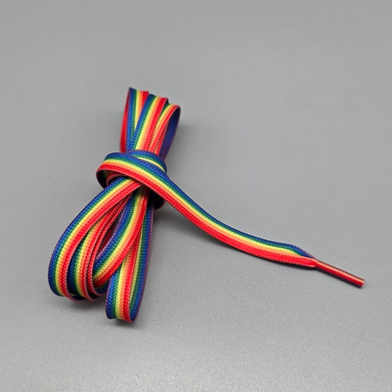 Ein flacher Schnürsenkel in Regenbogenfarben, der die horizontalen Streifen der Pride-Flagge zeigt: Rot, Orange, Gelb, Grün, Blau und Violett. Der Schnürsenkel ist links im Bild zu einem lockeren Bündel aufgewickelt, während ein Ende sich diagonal nach rechts unten erstreckt und in einer passenden farbigen Spitze (Aglet) endet. Der Hintergrund ist ein schlichter, gleichmäßiger Grauton.