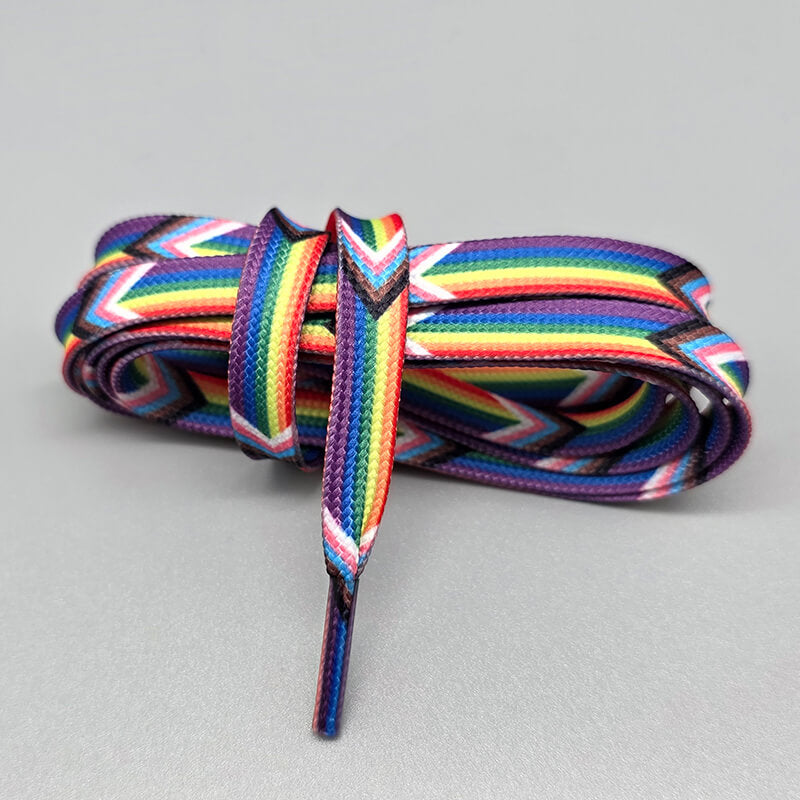 Nahaufnahme eines Paars aufgerollter Pride-Schnürsenkel der Variante "Rainbow Progress Classic". Die flachen, gewebten Schnürsenkel kombinieren die horizontalen Streifen der Regenbogenflagge mit dem Chevron-Muster der Progress-Pride-Flagge (Weiß, Rosa, Hellblau, Braun und Schwarz). Das Bündel liegt auf einem schlichten, grauen Hintergrund.