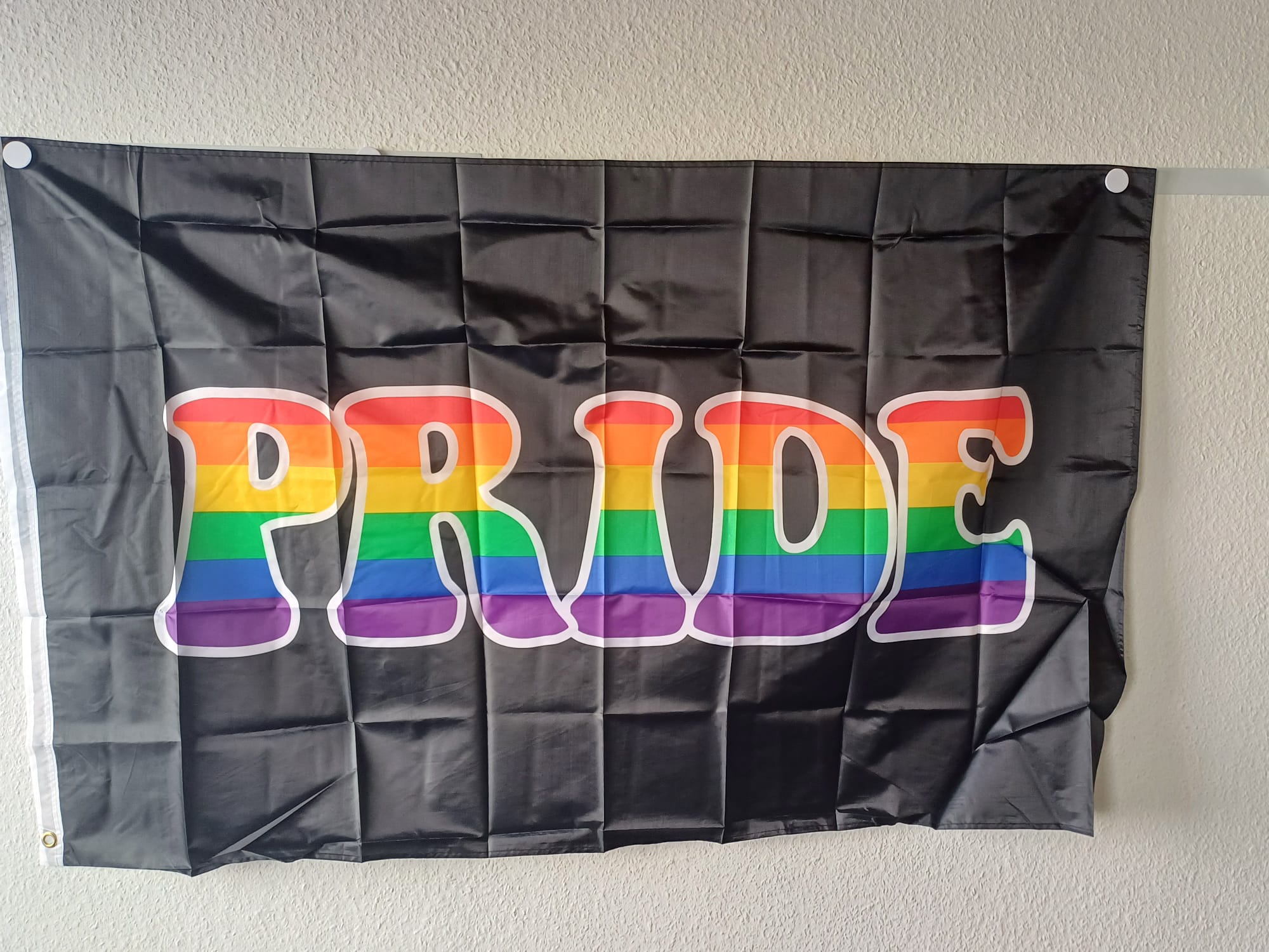 Die schwarze PRIDE-Flagge hängt ausgebreitet an einer hellen Wand. Der große, zentrale Schriftzug ist in den sechs Farben des Regenbogens ausgefüllt, weiß umrandet und links an der verstärkten weißen Kante sind die Befestigungsösen sichtbar.