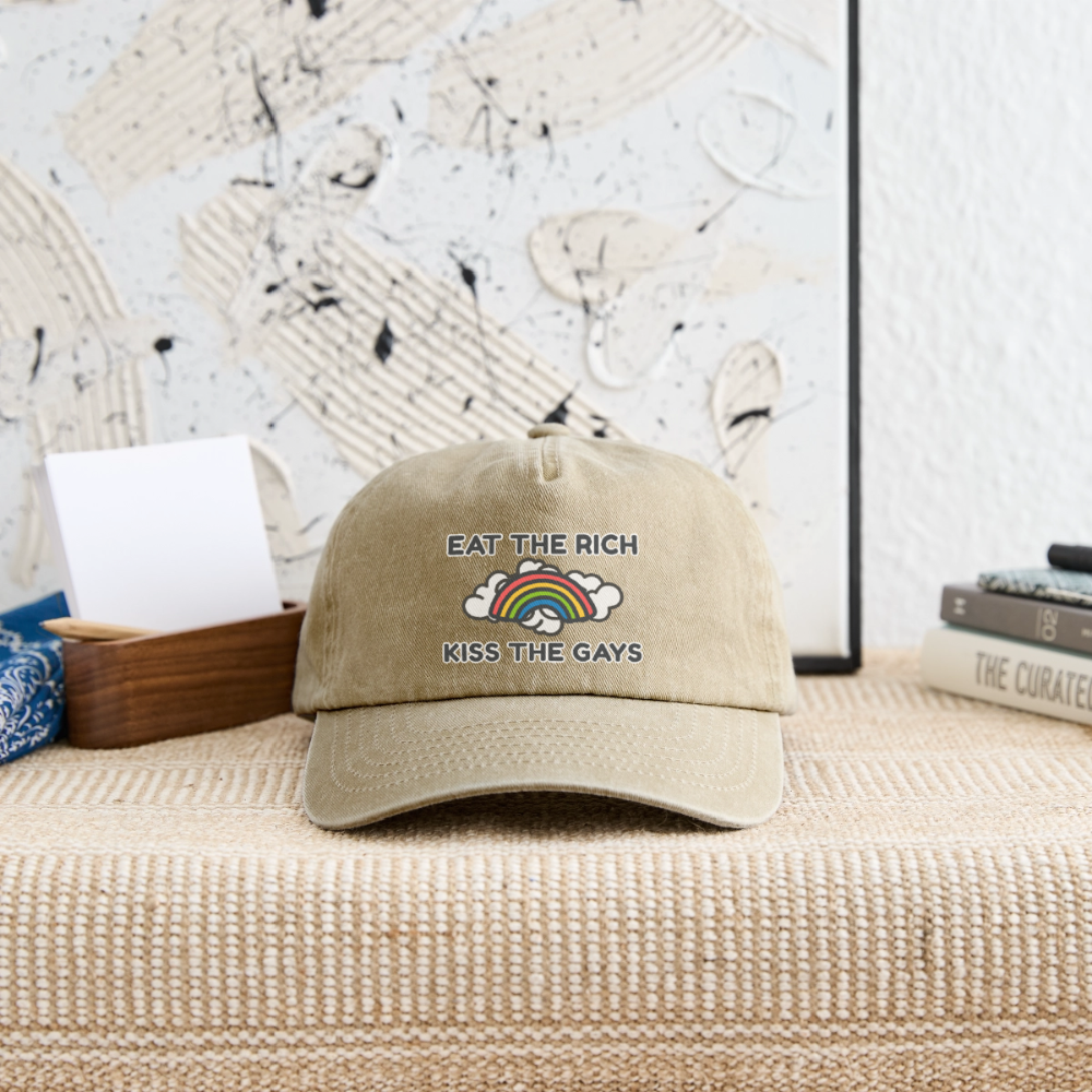 Stilvoll in Szene gesetzte beige Vintage Cap mit dem provokanten Spruch „Eat The Rich Kiss The Gays“. Der bunte Regenbogen verbindet harte politische Ansage mit queerer Leichtigkeit, abgelegt vor eleganter Kulisse.