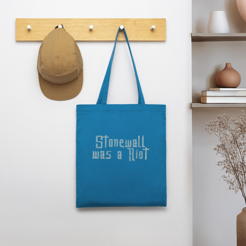Der königsblaue Tote Bag hängt an einer Holzgarderobe neben einer beigen Cap. Die raue Schablonenschrift auf der Tasche erinnert im heimischen Flur unübersehbar an den mutigen LGBTQ-Widerstand von 1969.