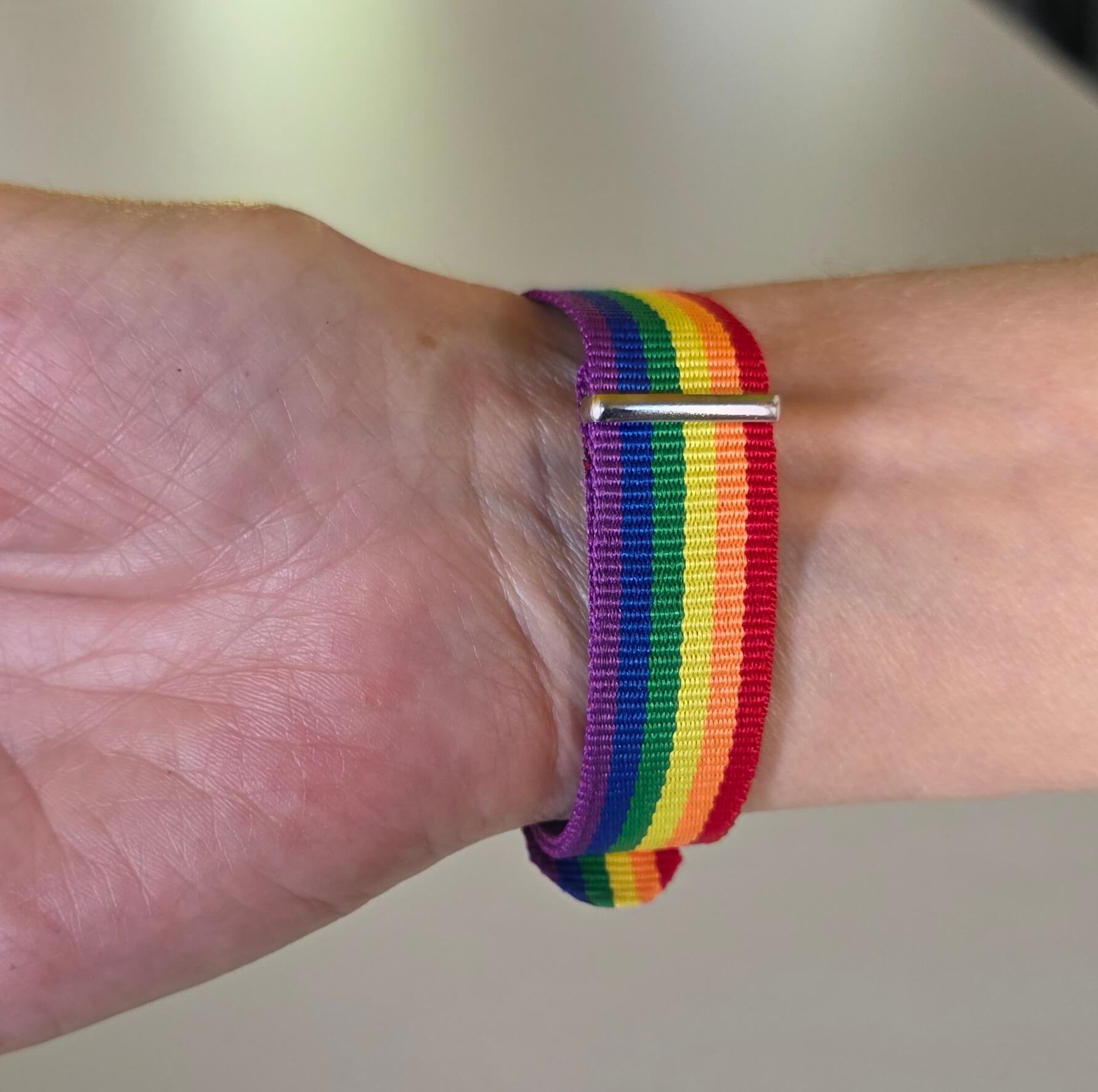 Eine Nahaufnahme zeigt das Regenbogen-Armband direkt an einem Unterarm getragen. Der Blick fällt genau auf die silberfarbene Metallschnalle. Der kleine Dorn steckt sicher in einem der gestanzten Löcher des Nylonbandes, während das farbenfrohe, überstehende Ende sauber durch die rechteckige Metallschlaufe geführt ist.