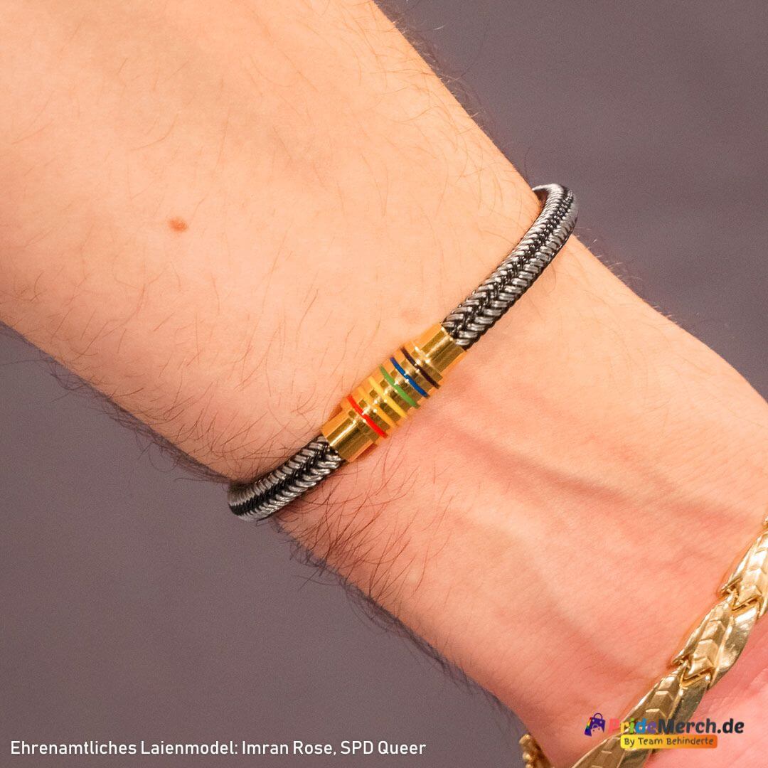 Präsentation des hochglanzpolierten Edelstahl-Armbands an dem Laienmodell Imran Rose. Die Nahaufnahme am Holztisch fokussiert das Handgelenk und hebt die leuchtenden Pride-Farben des Schmuckstücks im Kontrast zur dunklen Kleidung hervor.