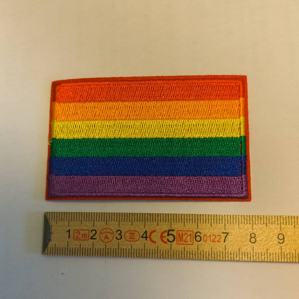 Ein dicht gestickter, rechteckiger Aufnäher mit den sechs horizontalen Streifen der klassischen Regenbogenflagge (Rot, Orange, Gelb, Grün, Blau, Violett). Der Patch liegt flach auf einem hellen Untergrund, direkt neben einem gelben Zollstock aus Holz zur Größendarstellung. Die sauber umkettelten Ränder und die strukturierte Stickerei sind deutlich erkennbar.