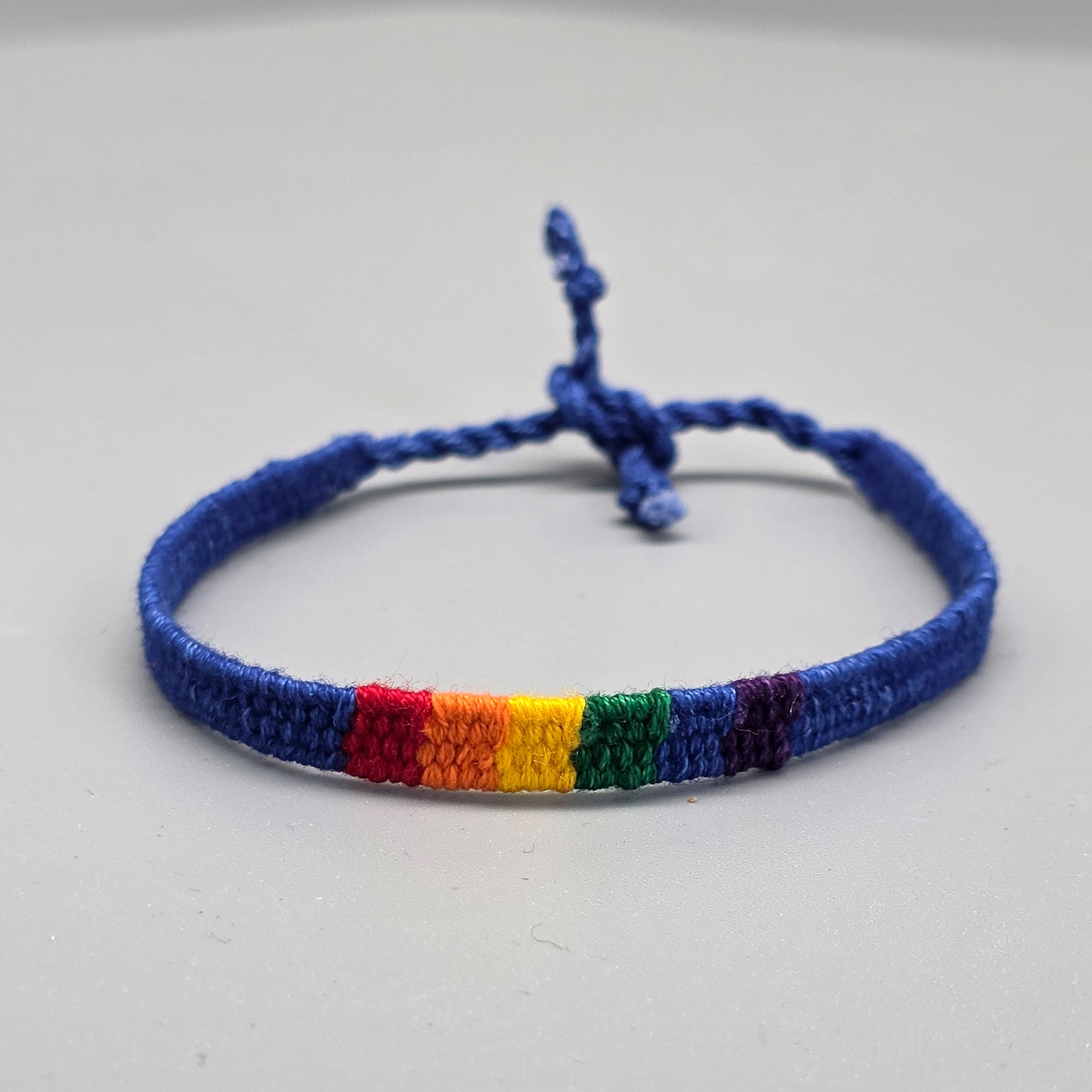 Ein handgemachtes, blaues Freundschaftsarmband aus geknoteter Baumwolle liegt in runder Form auf grauem Untergrund. Die Vorderseite präsentiert ein dezent eingewebtes Rechteck in den sechs klassischen Regenbogenfarben der Pride-Bewegung.