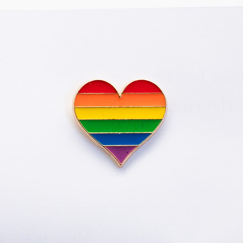 Ein herzförmiger Emaille-Pin liegt flach auf einer hellen Holzoberfläche und zeigt die sechs kräftigen Querstreifen der klassischen LGBTIQ-Regenbogenflagge. Das farbintensive Motiv wird von einer eleganten, silberfarbenen Metallkontur eingefasst.