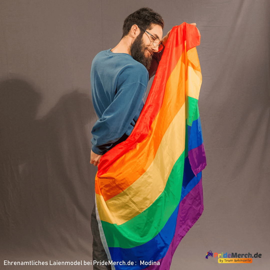 Laienmodell Modina ist dem Betrachter leicht abgewandt und blickt über seine rechte Schulter zurück in die Kamera. Die Regenbogenflagge verdeckt beinahe seinen gesamten Rücken und leuchtet intensiv vor dem grauen Stoffhintergrund. Er hält die Flagge mit einer Hand an seiner Schulter fest und schmunzelt leicht.