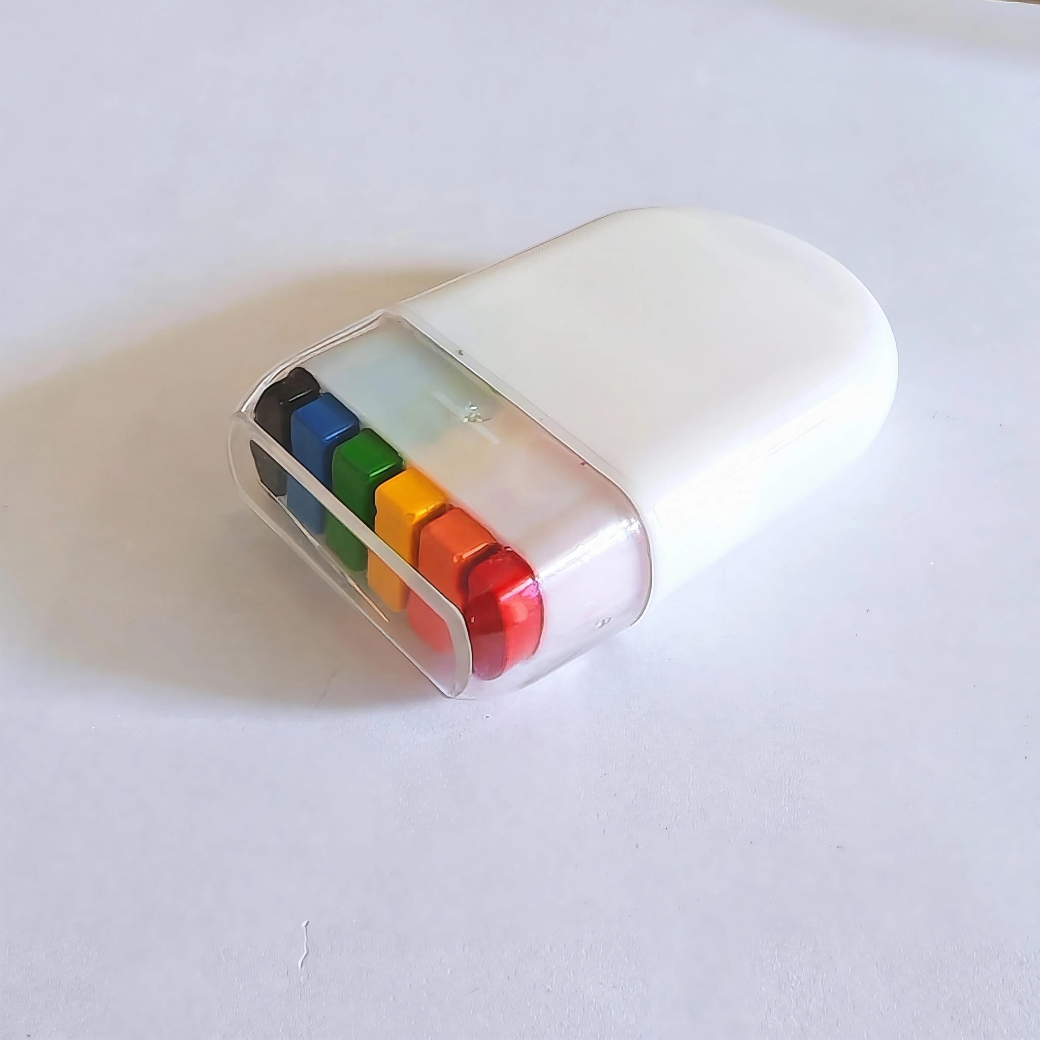 Der kompakte, weiße Kunststoff-Schminkstift liegt geschlossen auf einem weißen Untergrund. Die transparente Schutzkappe ist fest aufgesteckt. Durch die durchsichtige Kappe leuchten die sechs Wachsfarben in Schwarz, Blau, Grün, Gelb, Orange und Rot gut geschützt hindurch.