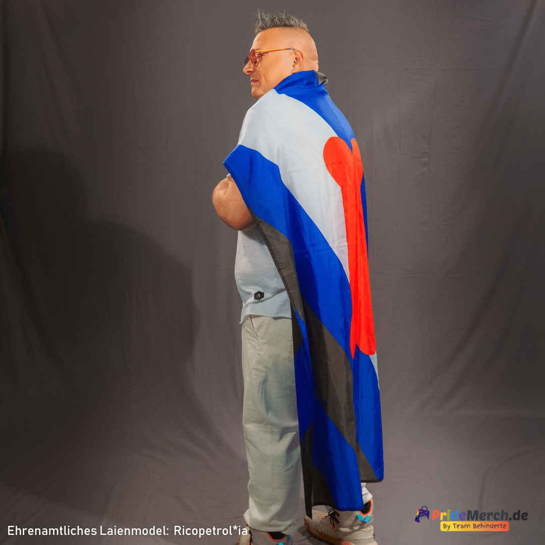 aienmodell Ricopetrol*ia trägt die blaue, schwarze und weiße Puppy Play Flagge wie einen Umhang über den Schultern. Die Seitenansicht zeigt den dekorativen Faltenwurf des leichten Polyesterstoffs im Fotostudio vor dunklem Hintergrund.