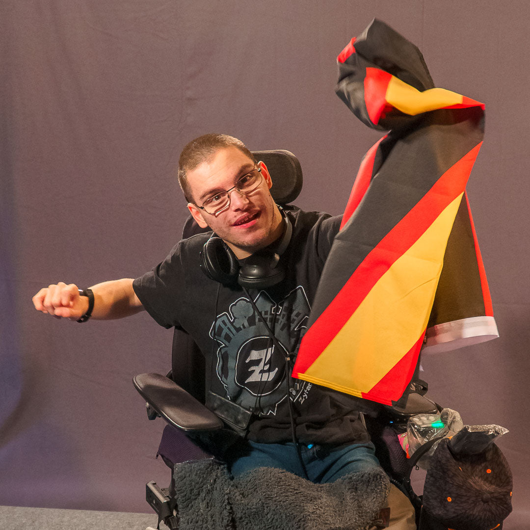 Model Sandro Weigang sitzt in seinem Rollstuhl vor einem grauen Fotohintergrund. Er trägt eine Brille, ein dunkles T-Shirt und schwarze Kopfhörer um den Hals. Sandro lächelt und hält die Rubber Pride Flagge mit der linken Hand dynamisch nach oben. Die Flagge zeigt einen schwarzen Hintergrund mit einem gelb-roten, nach unten abgewinkelten Streifen.