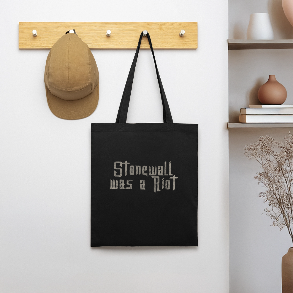 Neben einer Basecap an einer Holzgarderobe hängt der schwarze PrideLAB-Beutel. Die helle, markante Stencil-Typografie fällt im Raum sofort ins Auge und trägt den Aktivismus direkt auf die Straße.