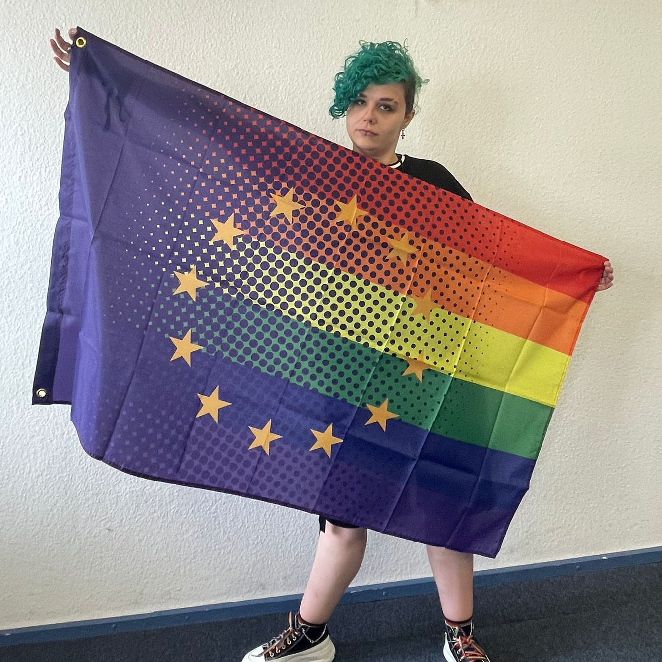 Das ehrenamtliche Laienmodell Shay Heemsoth-Ohlmeier (Mitorganisatorin des CSD Verden) hält die Regenbogen Europa Pride Flagge weit ausgebreitet vor einer weißen Wand. Das Foto zeigt die europäische Flagge mit integriertem Halfton-Punktraster auf den sechs queeren Regenbogenstreifen in voller Pracht.