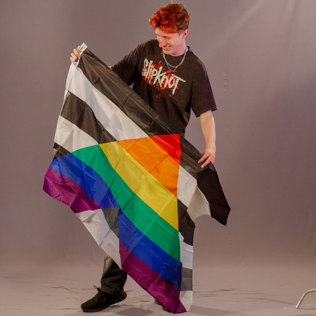 Das Laienmodell blickt mit einem breiten Lächeln schräg nach unten. Er trägt ein schwarzes Band-Shirt und dunkle Hosen. Er hält die Straight Ally Flagge so vor seinem Körper, dass der Stoff leicht in Falten fällt. Das leuchtende, regenbogenfarbene „A“ auf dem schwarz-weißen Streifenhintergrund sticht deutlich als zentrales Motiv hervor.