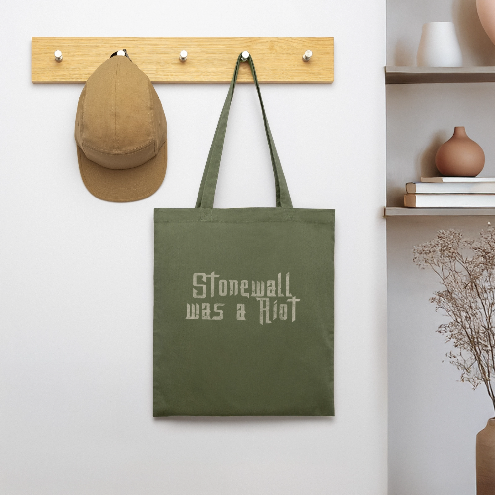 An einem Holz-Kleiderhaken hängt die khakifarbene Pride-Tasche. Die raue Typografie auf dem Stoff setzt in der hellen Raumumgebung ein unmissverständliches politisches Zeichen für echten Aktivismus.