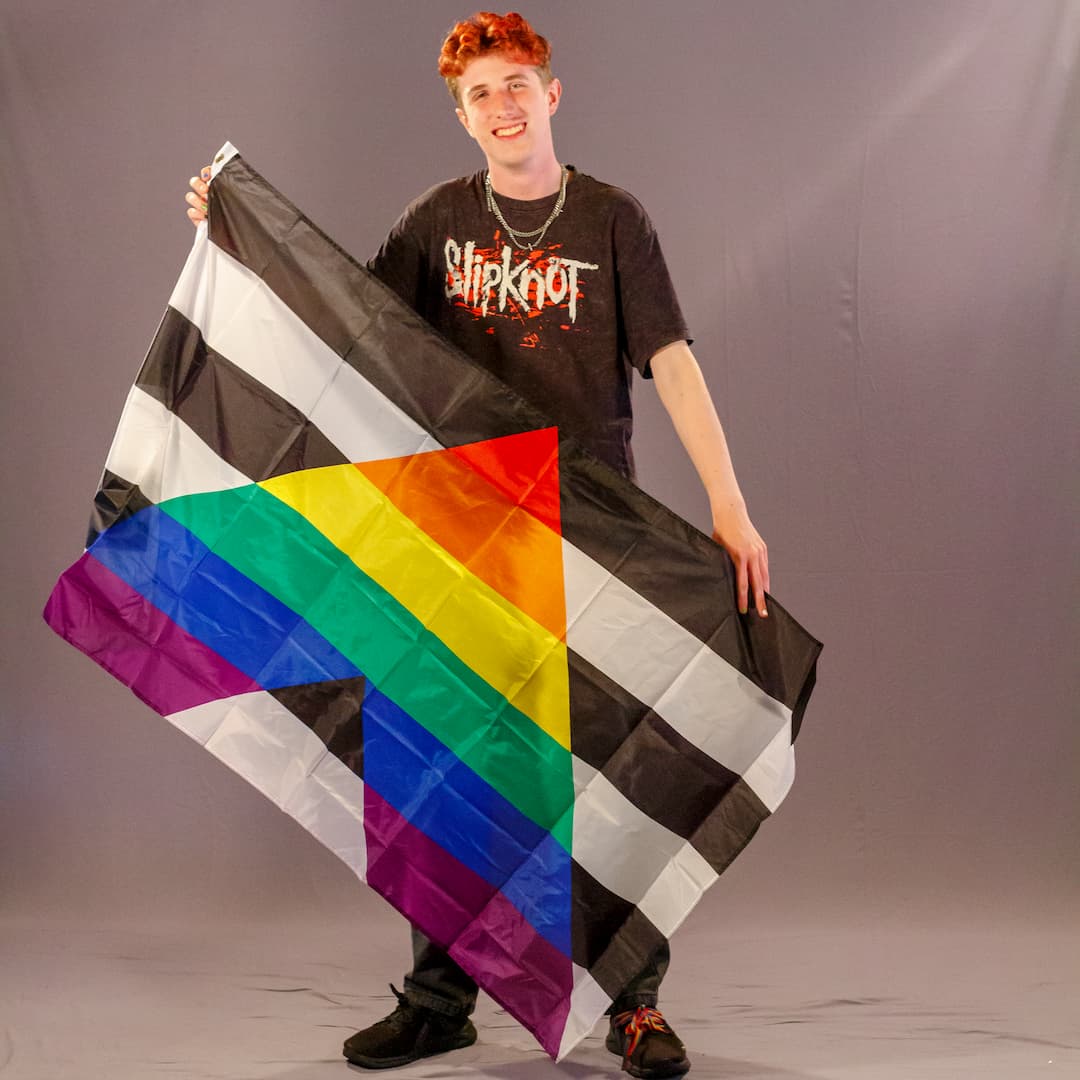 Ein Laienmodell mit rötlichen Haaren und schwarzen Sneakern mit bunten Schnürsenkeln, blickt direkt und freudig in die Kamera. Er breitet die Straight Ally Flagge großflächig vor seinen Beinen und seinem Oberkörper aus. Das kontrastreiche Design – schwarz-weiße Querstreifen durchbrochen von einem großen Regenbogen-Dreieck – ist in seiner vollen Breite gut zu erkennen.