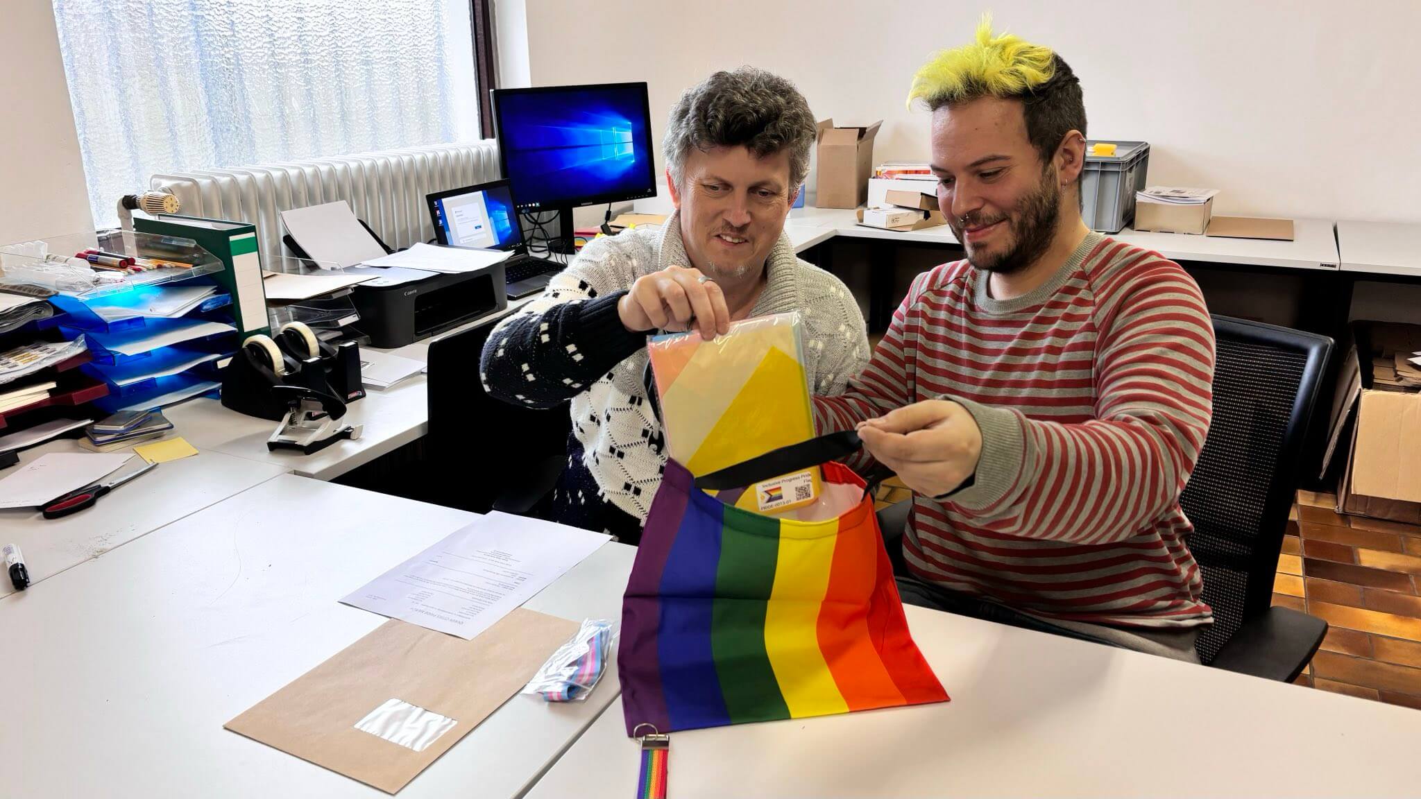 Thorsten Höller, der Initiator des Team Behinderte vom Queer Cities e.V., und sein Kollege Passilo Biermanski arbeiten gemeinsam an einem Schreibtisch im Büro von PrideMerch. Thorsten Höller packt konzentriert eine verpackte Regenbogenflagge in eine bunte Regenbogen-Tasche, die Passilo Biermanski, der markante neongelbe Haare hat und lächelt, offen hält. Auf dem Arbeitstisch liegen Versandunterlagen und ein Regenbogen-Schlüsselband. Im Hintergrund ist ein Büro mit Computer-Arbeitsplätzen. Gelebte Inklusion.