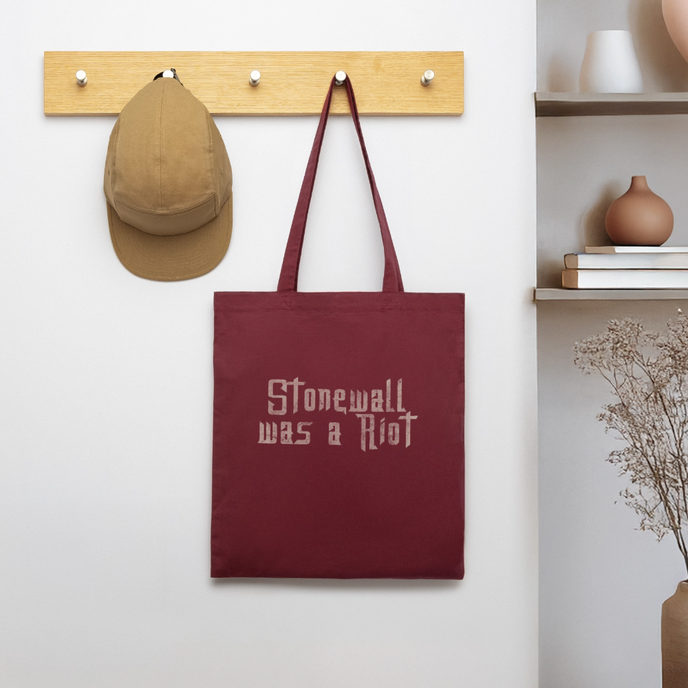Hängend an einer Holzleiste präsentiert sich der burgunderrote Beutel als täglicher Begleiter. Der helle Used-Look-Schriftzug erinnert neben dem Regal mit Vasen subtil, aber deutlich an die radikalen Wurzeln der queeren Befreiung.