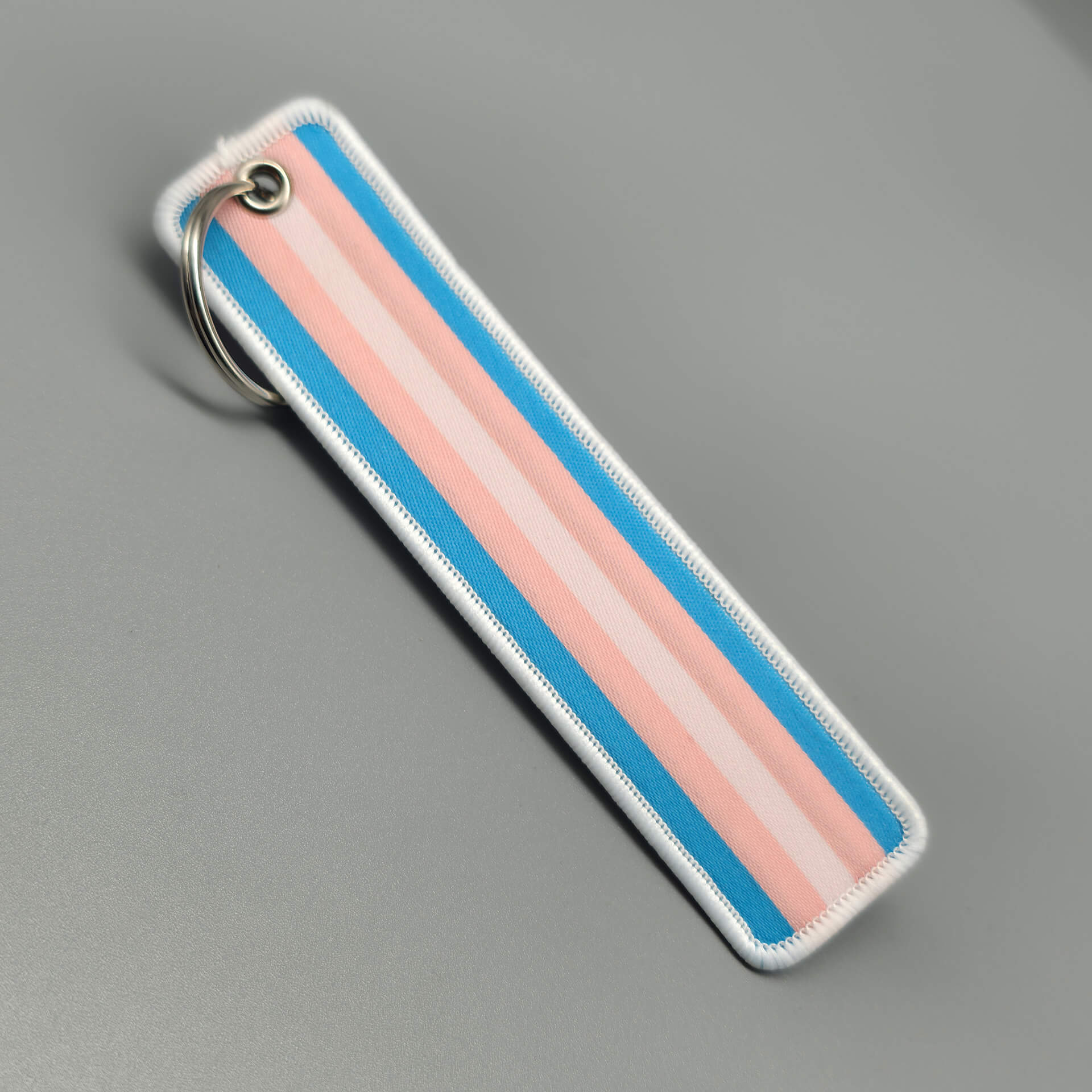 Ein einzelner Schlüsselanhänger aus fest gewebtem Textil liegt diagonal auf einer grauen Fläche. Das Design zeigt die horizontalen Streifen der Trans-Flagge in Hellblau, Rosa und Weiß. Eine dicke weiße Kettelnaht schützt die Ränder, während am oberen Ende ein silberfarbener Metallring durch eine Öse befestigt ist. Ein subtiles Statement für geschlechtliche Vielfalt.