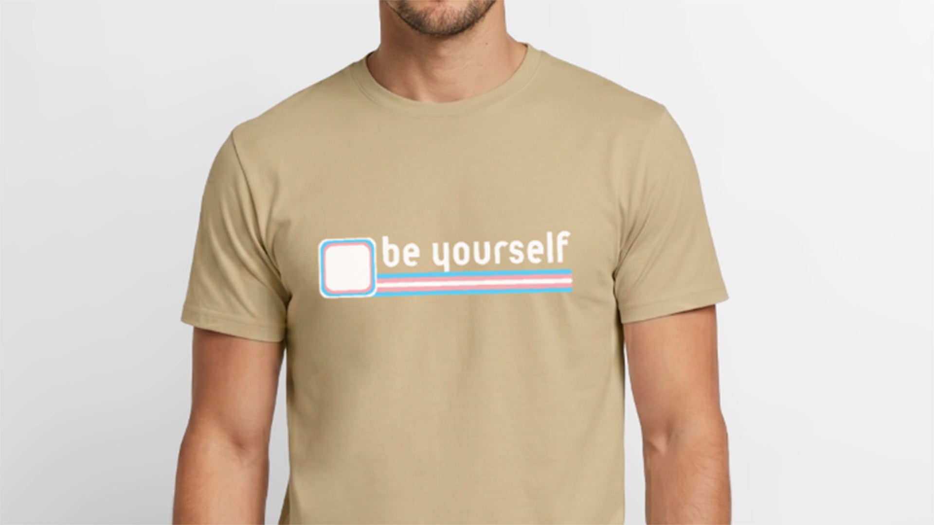 Nahaufnahme eines Models im sandbeigen T-Shirt mit dem starken Statement „be yourself". Die präzisen Linien der Trans-Flagge in Hellblau, Rosa und Weiß fordern unmissverständlich zu echter Sichtbarkeit auf – ein Banner-Motiv, das für kompromisslosen Pride steht.