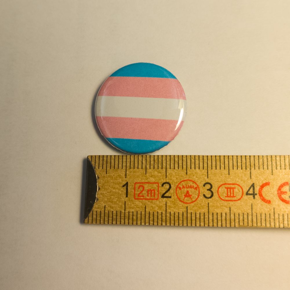 Runder Metall-Button mit den fünf Querstreifen der Transgender-Flagge in Blau, Pink und Weiß. Zur Größendarstellung liegt der Anstecker neben einem hölzernen Zollstock, der auf den Bildern einen Durchmesser von 25 Millimetern anzeigt.

