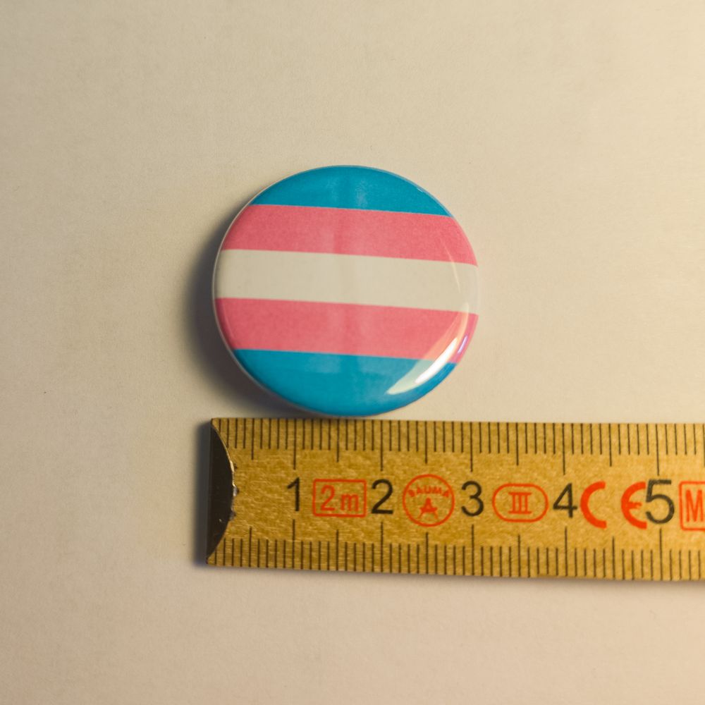 Fünf sanfte Querstreifen in Hellblau, Rosa und Weiß zieren die glänzende Oberfläche dieses Ansteckers und zeigen die klassische Transgender-Flagge. Direkt unter dem Schmuckstück liegt ein gelber Holz-Zollstock, an dem sich ein Durchmesser von knapp 31 Millimetern ablesen lässt.