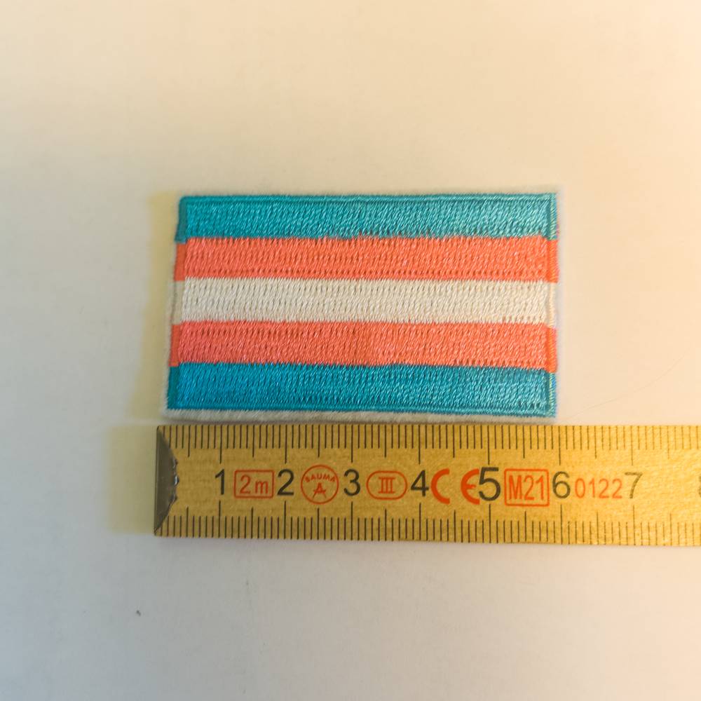 Ein sauber umrandeter, gestickter Patch mit den fünf horizontalen Streifen der Transgender-Flagge: Hellblau, Rosa, Weiß, Rosa, Hellblau. Das Produkt liegt flach auf, direkt unterhalb des Aufnähers liegt ein gelber Zollstock, an dem man die exakte Breite ablesen kann.