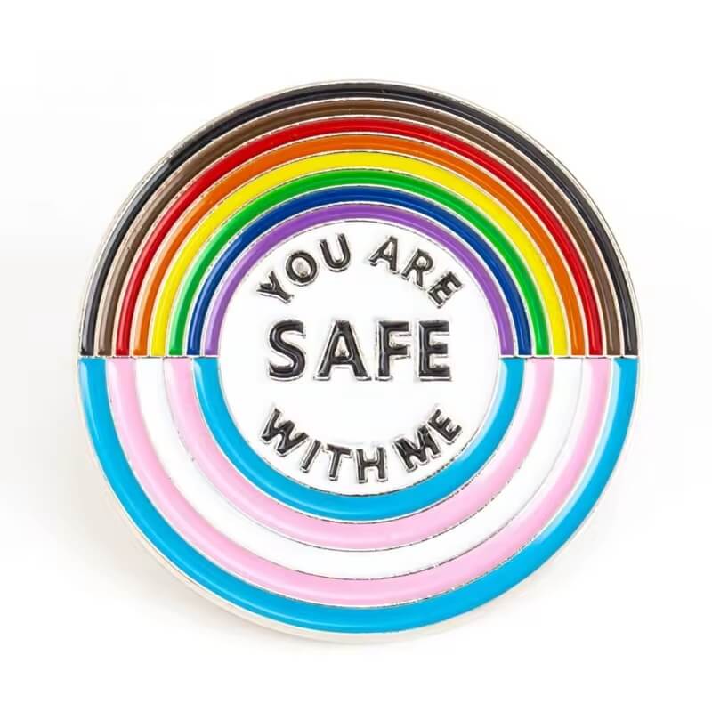 Runder Ansteck-Pin mit der Aufschrift „YOU ARE SAFE WITH ME“ in der Mitte. Umgeben von Halbkreisen in den Farben der Progress Pride Flag: Braun, Schwarz, Rot, Orange, Gelb, Grün, Blau, Lila, sowie die Farben der Trans*-Flagge in Rosa, Blau und Weiß. Symbol für Solidarität, Sicherheit und Zugehörigkeit zur LGBTQIA+ Community.