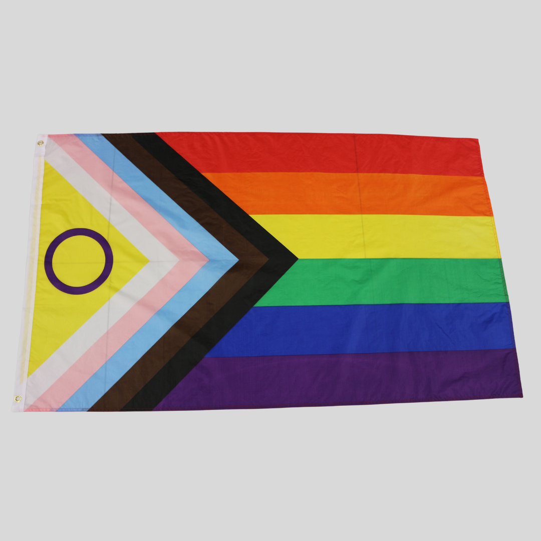 Die Intersex-Inclusive-Progress-Pride-Flagge liegt ausgebreitet auf einer dunklen Holzoberfläche und zeigt das vollständige, erweiterte Farbschema. Links im Keil sind die hellblau-rosa-weißen Trans-Farben sowie das gelbe Dreieck mit dem lila Kreis zu sehen.