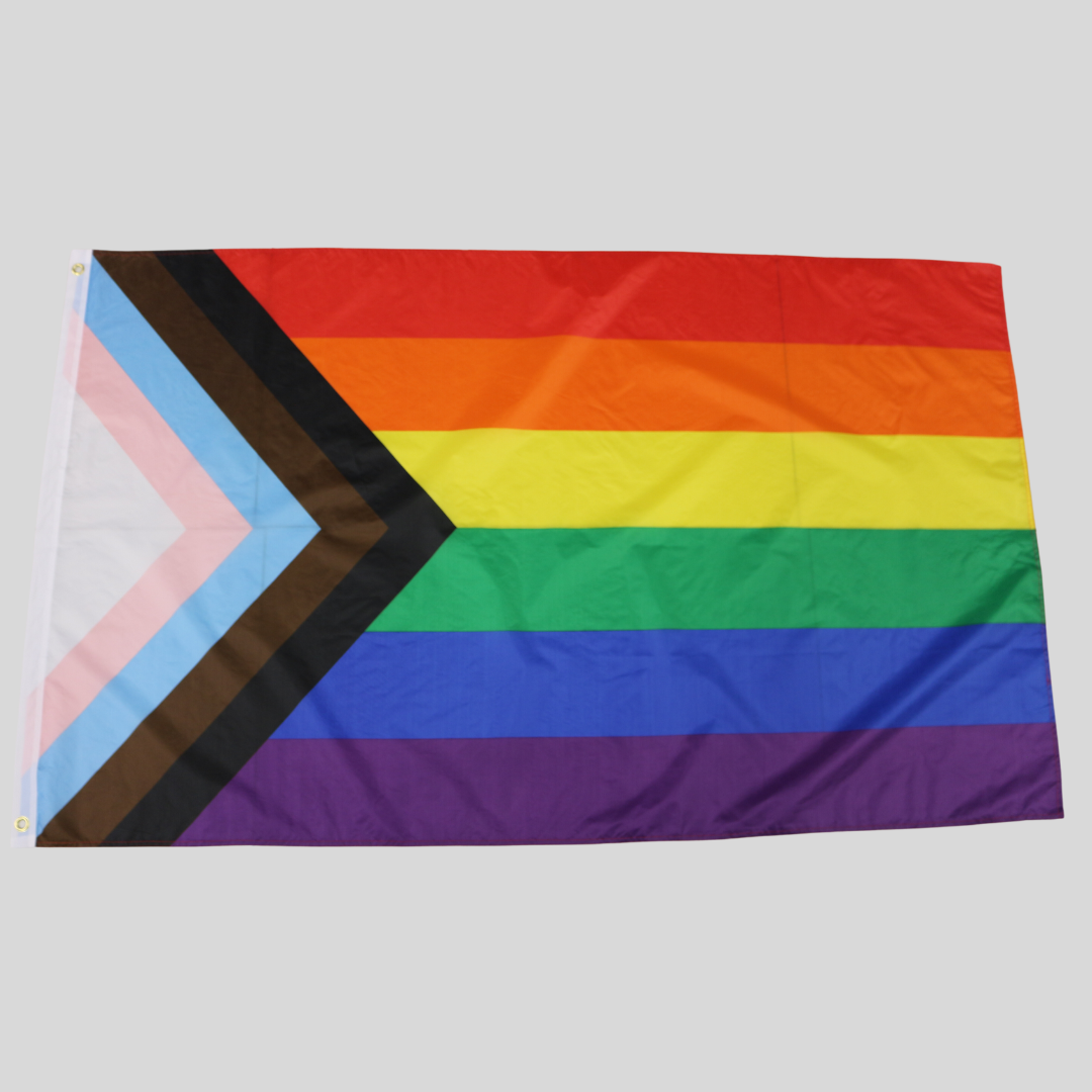 Eine flach ausgebreitete Progress Classic Pride Flagge vor einem hellgrauen Hintergrund. Auf der rechten Seite verlaufen die sechs klassischen Regenbogenstreifen waagerecht. Von links schiebt sich ein keilförmiges Pfeilmotiv in den Regenbogen, bestehend aus den Farben Weiß, Rosa, Hellblau, Braun und Schwarz. An der linken, weißen Stoffkante sind zwei messingfarbene Metallösen zur Befestigung zu sehen.
