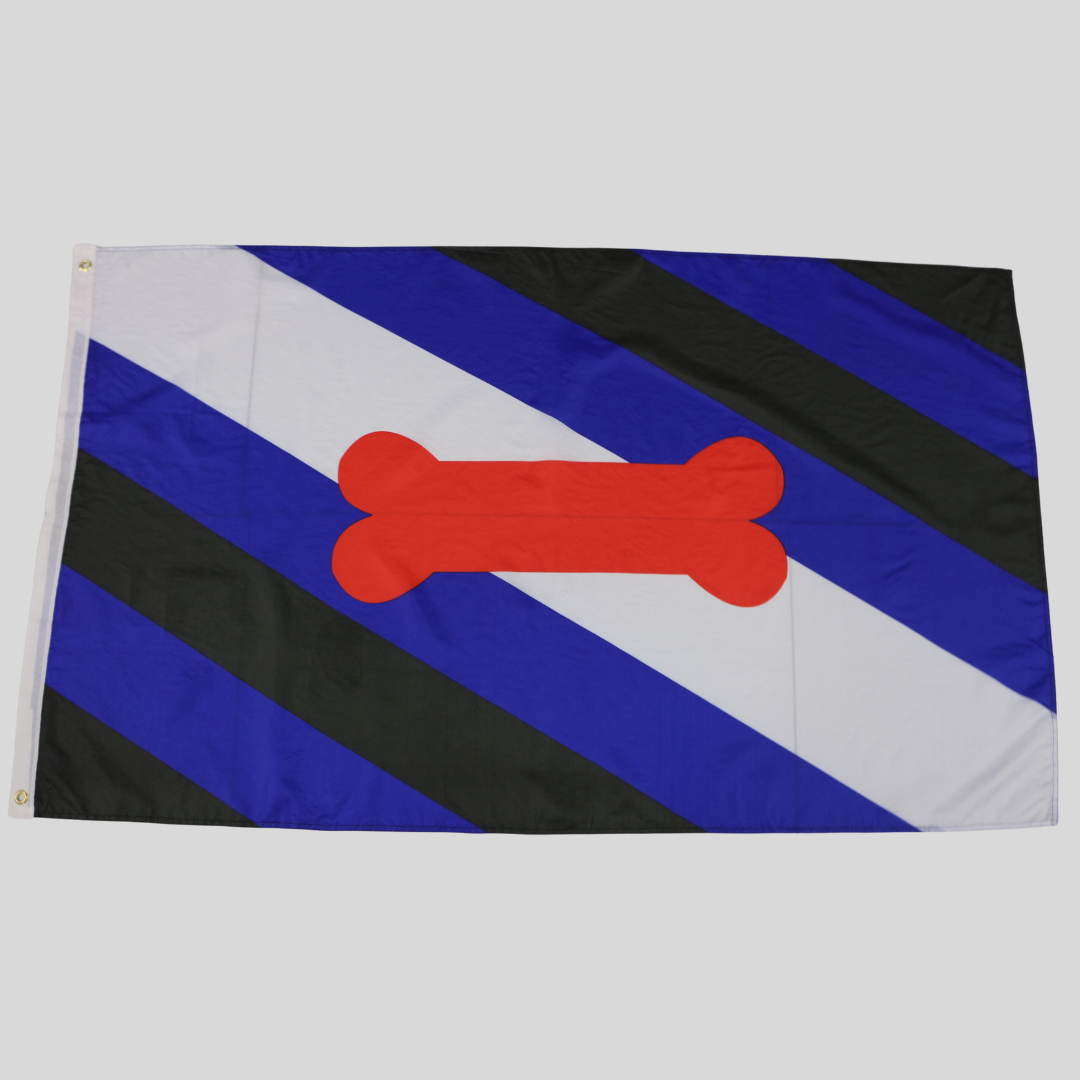 Die Puppy Play Pride Flagge liegt komplett flach ausgebreitet auf einer neutralen grauen Studiofläche. Gut sichtbar sind die diagonalen Streifenverläufe, der rote Knochen sowie das weiße Besatzband mit den zwei Metallösen zur Befestigung auf der linken Seite.